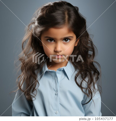 Portrait of an angry little girl with wavy brown hair. Closeup face of a furious Caucasian child on a blue background. White European small kid with no expression in a blue shirt looking at camera. 105437177