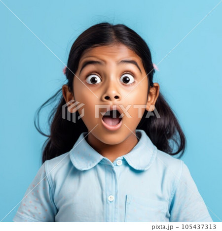 Portrait of a surprised little Indian girl with big eyes and an open mouth. Closeup face of an amazed Indonesian child on a blue background. Astonished kid in a blue shirt looking at the camera. 105437313