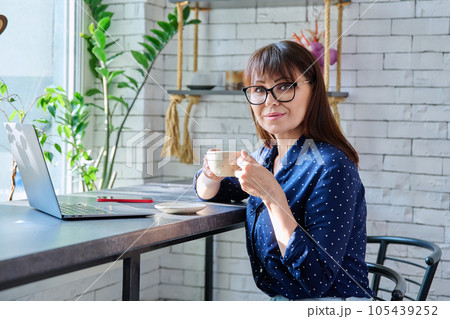 Middle-aged woman at coffee shop drinking coffee, with laptop, looking at camera Middle-aged woman at coffee shop drinking coffee, with laptop, looking at camera 105439252