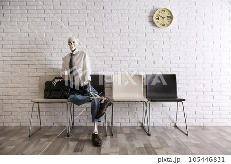 Human skeleton in office wear sitting on chair near brick wall indoors 105446831