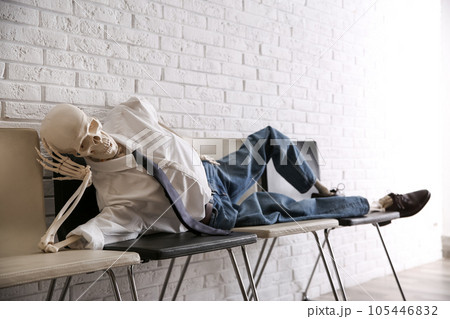Human skeleton in office wear lying on chairs near brick wall indoors 105446832