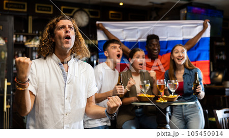 Cheerful international soccer football fans waving Russian flag and drinking beer, eating chips in the sport bar 105450914