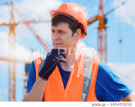 Portrait of pensive builder against sky with tower cranes. Builder handyman in hard hat stands with Portrait of pensive builder against sky with tower cranes. Builder handyman in hard hat stands with 105455010