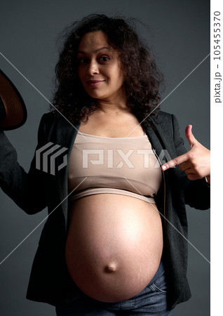 Cheerful pregnant woman pointing her finger on her pregnant belly, smiling looking at camera, isolated studio background 105455370