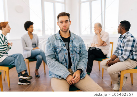 Portrait of sad Caucasian man looking at camera with serious expression while group of multicultural and different ages people sitting in circle, talking in background during therapy session. Portrait of sad Caucasian man looking at camera with serious expression while group of multicultural and different ages people sitting in circle, talking in background during therapy session. 105462557