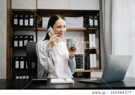 Business Caucasian woman Talking on the phone and using a laptop with a smile while sitting at modern office Business Caucasian woman Talking on the phone and using a laptop with a smile while sitting at modern office 105464377