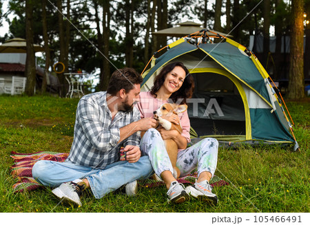 Young married couple with their corgi dog sitting in the forest near tent and looking at sunset 105466491