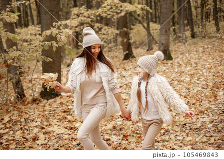 Young woman with little girl walking in autumn forest. Brunette woman play with her daughter. Girl wearing beige sweater and mother wearing white clothes. 105469843