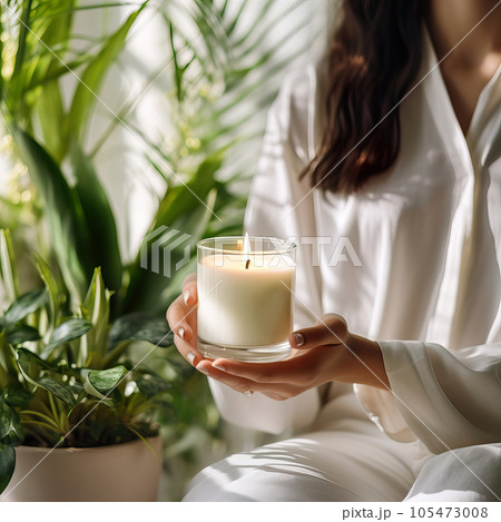Young woman holding burning candle jar in her hands, container candle mockup closeup shot, mindfulness home interior with green plants 105473008