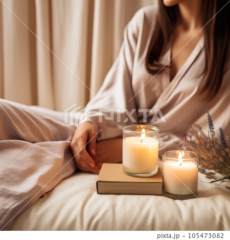 Beautiful young woman with long hair dressed in white relaxing in her home bedroom with burning candles, flowers and books, cozy home decor in natural colors. Candle jar mockup 105473082