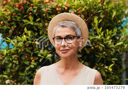 Portrait of a stylish mature woman. Senior female with gray hair wearing glasses and a small straw hat.	 105473279