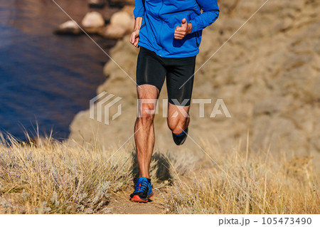 front view runner running mountain uphill in blue jacket and black tights, trail dry grass front view runner running mountain uphill in blue jacket and black tights, trail dry grass 105473490
