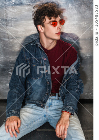 Portrait of trendy stylish attractive fashion model man in bright red sunglasses and denim casual style sitting on floor near metallic wall and looking away. Indoor studio shot. 105485583
