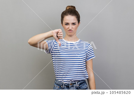 Portrait of unhappy unsatisfied young adult woman wearing striped T-shirt showing thumb down, dislike gesture, negative emotions, bad rating. Indoor studio shot isolated on gray background. Portrait of unhappy unsatisfied young adult woman wearing striped T-shirt showing thumb down, dislike gesture, negative emotions, bad rating. Indoor studio shot isolated on gray background. 105485620