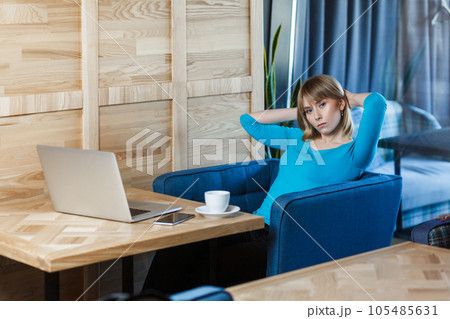 Portrait of serious self confident young woman with blonde hair in blue shirt sitting at table and working on laptop, looking at camera with bossy concentrated face. Indoor shot, cafe background. 105485631