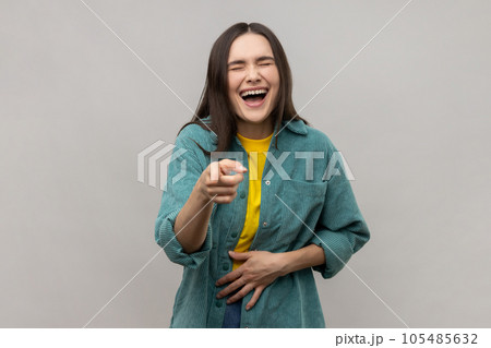 Joyful woman laughing, holding stomach, pointing to camera, taunting you, can't stop hysterical laughter, wearing casual style jacket. Indoor studio shot isolated on gray background. 105485632