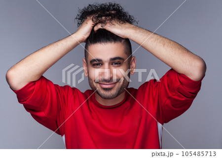 Portrait of handsome attractive smiling bearded man with curly hair, wearing red jumper, standing looking at camera, collected his hair. Indoor studio shot isolated on gray background. Portrait of handsome attractive smiling bearded man with curly hair, wearing red jumper, standing looking at camera, collected his hair. Indoor studio shot isolated on gray background. 105485713