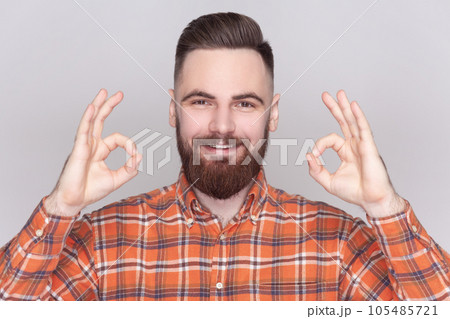 Smiling cheerful joyful bearded man standing looking at camera, showing okay sign, ok gesture, expressing positive emotions, wearing checkered shirt. Indoor studio shot isolated on gray background. Smiling cheerful joyful bearded man standing looking at camera, showing okay sign, ok gesture, expressing positive emotions, wearing checkered shirt. Indoor studio shot isolated on gray background. 105485721