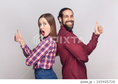 Portrait of young adult woman and man standing back to back together showing thumbs up, like gesture, positive feedback, looking at camera. Indoor studio shot isolated on gray background. 105485726