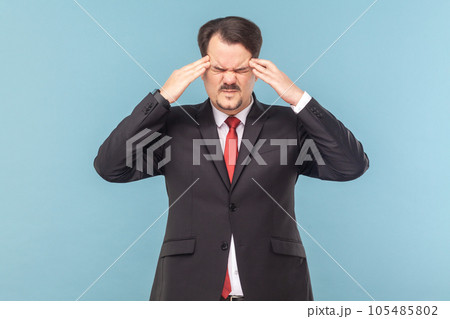 Sick ill man with mustache standing feels unwell, suffering headache, frowning face, massaging temples, wearing black suit with red tie. Indoor studio shot isolated on light blue background. 105485802