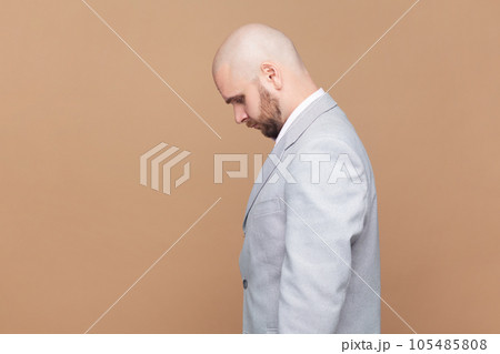 Side view portrait of sad upset bald bearded man standing with head down, being offended, expressing sorrow and sadness, wearing gray jacket. Indoor studio shot isolated on brown background. Side view portrait of sad upset bald bearded man standing with head down, being offended, expressing sorrow and sadness, wearing gray jacket. Indoor studio shot isolated on brown background. 105485808