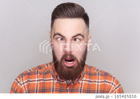 Portrait of comic positive bearded man looking cross-eyed, having fun with silly face expression, playing fool, wearing checkered shirt. Indoor studio shot isolated on gray background. 105485810