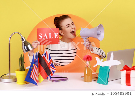 Woman employee of travel agency sitting in rubber ring on neck, showing sale card and screaming in megaphone, hot tours to different countries. Indoor studio studio shot isolated on yellow background. 105485901