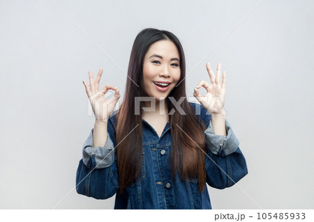 Portrait of optimistic positive young adult brunette woman in blue denim jacket standing showing okay sign, looking smiling at camera, Indoor studio shot isolated on gray background. Portrait of optimistic positive young adult brunette woman in blue denim jacket standing showing okay sign, looking smiling at camera, Indoor studio shot isolated on gray background. 105485933