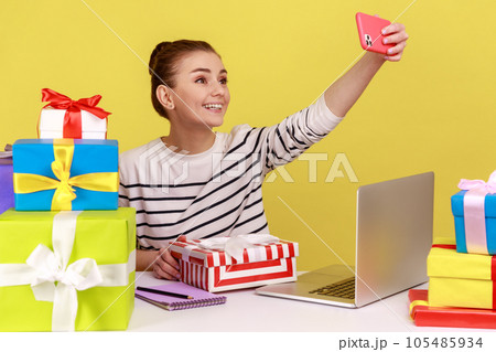 Friendly woman making selfie on smartphone camera sitting at workplace with many colorful gift boxes around, holidays or birthday celebration. Indoor studio studio shot isolated on yellow background. Friendly woman making selfie on smartphone camera sitting at workplace with many colorful gift boxes around, holidays or birthday celebration. Indoor studio studio shot isolated on yellow background. 105485934