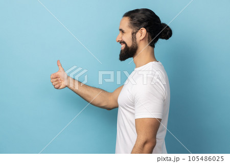 Side view of man with beard wearing white T-shirt standing with thumbs up, like gesture, demonstrating approval and agree with suggestion. Indoor studio shot isolated on blue background. 105486025