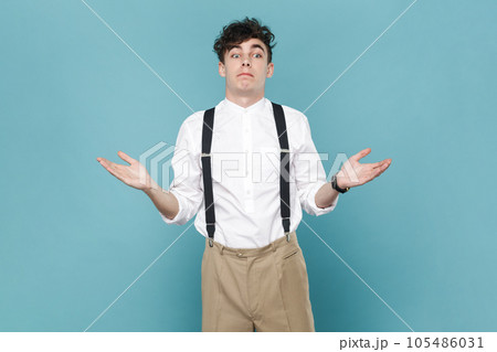 Portrait of puzzled confused man wearing white shirt and suspender, shrugging shoulders, looking away, raised his arms, has uncertain expression. Indoor studio shot isolated on blue background. 105486031