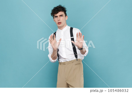 Portrait of scared frighten man wearing white shirt and suspender trying to protect himself from danger, showing stop gesture with palms. Indoor studio shot isolated on blue background. Portrait of scared frighten man wearing white shirt and suspender trying to protect himself from danger, showing stop gesture with palms. Indoor studio shot isolated on blue background. 105486032