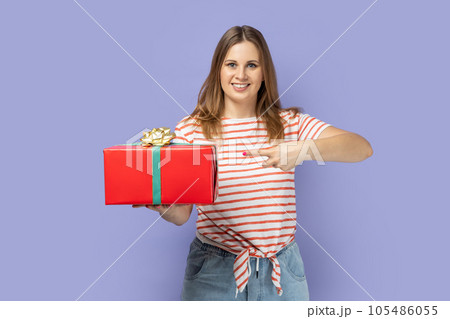 Portrait of pretty good looking attractive blond woman wearing striped T-shirt holding and pointing finger at red present box, smiling to camera. Indoor studio shot isolated on purple background. 105486055