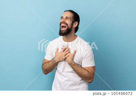 Portrait of overjoyed cheerful man with beard wearing white T-shirt keeps hands on chest closes eyes and expressing positive emotions. Indoor studio shot isolated on blue background. Portrait of overjoyed cheerful man with beard wearing white T-shirt keeps hands on chest closes eyes and expressing positive emotions. Indoor studio shot isolated on blue background. 105486072