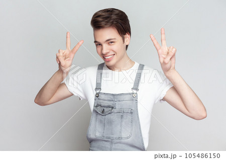 Portrait of young brunette man standing makes peace gesture, shows v sign, looking at camera with toothy smile, wearing denim overalls. Indoor studio shot isolated on gray background. Portrait of young brunette man standing makes peace gesture, shows v sign, looking at camera with toothy smile, wearing denim overalls. Indoor studio shot isolated on gray background. 105486150