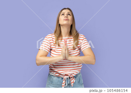 Portrait of attractive blond woman wearing striped T-shirt raising hands in prayer, looking up with hopeful pleading eyes, praying. Indoor studio shot isolated on purple background. Portrait of attractive blond woman wearing striped T-shirt raising hands in prayer, looking up with hopeful pleading eyes, praying. Indoor studio shot isolated on purple background. 105486154