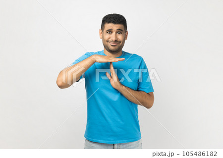 Portrait of attractive young adult unshaven man wearing blue T- shirt standing showing time out gesture with hands, looking at camera. Indoor studio shot isolated on gray background. 105486182