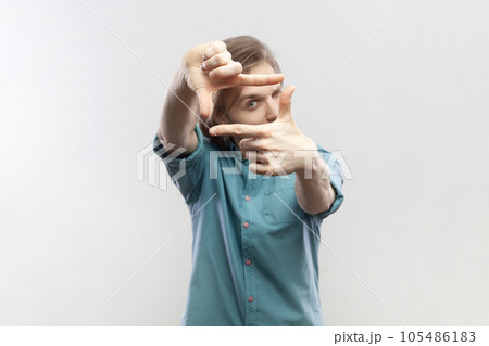 Portrait of man looking through frame of fingers gesturing photo, zooming focusing to camera, observing with serious expression, wearing blue shirt. Indoor studio shot isolated on gray background. 105486183