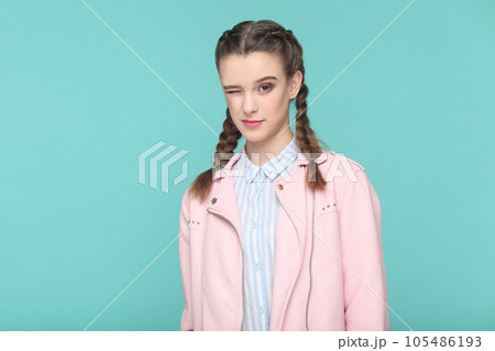 Portrait of playful cheerful teenager girl with braids wearing pink jacket standing looking at camera, winking, flirting with boyfriend. Indoor studio shot isolated on green background. Portrait of playful cheerful teenager girl with braids wearing pink jacket standing looking at camera, winking, flirting with boyfriend. Indoor studio shot isolated on green background. 105486193