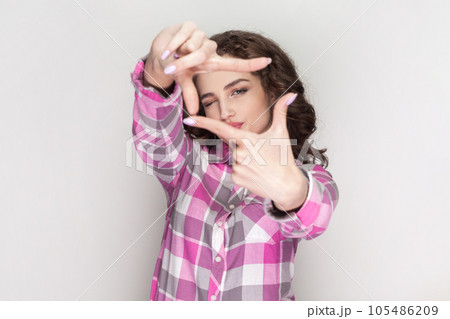 Happy ambitious woman with curly hair aims only success, makes hand frames searches perfect angle smiles broadly, gazes at camera through hands. Indoor studio shot isolated on gray background. 105486209