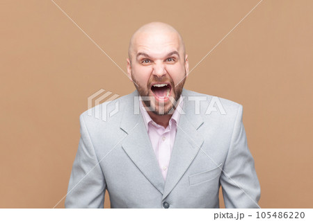 Portrait of angry or shocked bald bearded man standing, looking at camera and screaming, arguing with friend, wearing gray jacket. Indoor studio shot isolated on brown background. 105486220