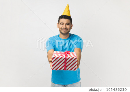 Portrait of cute attractive positive unshaven man wearing blue T- shirt and yellow party cone giving present box, having fun at birthday party. Indoor studio shot isolated on gray background. 105486230