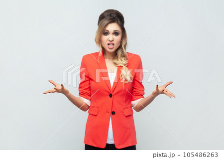 Portrait of angry irritated beautiful blonde woman standing with raised arms, screaming, asking why or what, arguing with somebody, wearing red jacket. Indoor studio shot isolated on gray background. 105486263