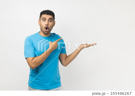 Portrait of amazed excited surprised astonished unshaven man wearing blue T- shirt standing presenting advertisement area on his palm, mockup. Indoor studio shot isolated on gray background. Portrait of amazed excited surprised astonished unshaven man wearing blue T- shirt standing presenting advertisement area on his palm, mockup. Indoor studio shot isolated on gray background. 105486267