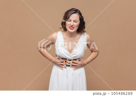 Portrait of unhealthy sick middle aged woman with wavy hair standing, holding her belly with hands, stomach cramps, wearing white dress. Indoor studio shot isolated on light brown background. 105486289