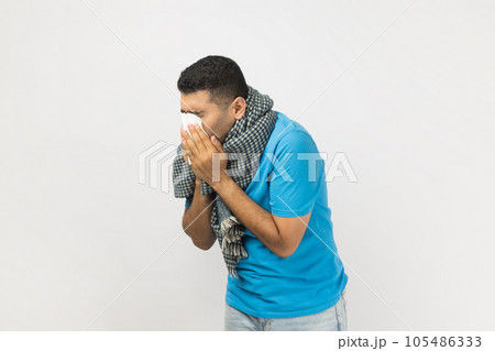 Portrait of ill sick unshaven man wearing blue T- shirt and wrapped in warm scarf standing sneezing, having runny nose, has grippe. Indoor studio shot isolated on gray background. Portrait of ill sick unshaven man wearing blue T- shirt and wrapped in warm scarf standing sneezing, having runny nose, has grippe. Indoor studio shot isolated on gray background. 105486333