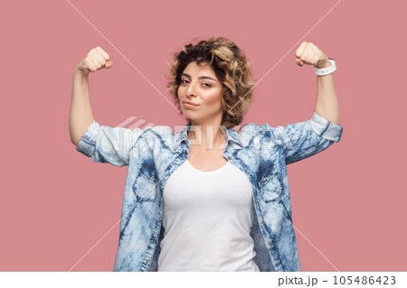 Portrait of self confident strong woman with curly hairstyle wearing blue shirt standing with raised arms, showing her power, looking at camera. Indoor studio shot isolated on pink background. Portrait of self confident strong woman with curly hairstyle wearing blue shirt standing with raised arms, showing her power, looking at camera. Indoor studio shot isolated on pink background. 105486423