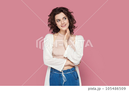 Portrait of friendly pleased smiling woman with curly hair wearing casual style outfit, looking away, dreaming about something pleasant. Indoor studio shot isolated on pink background. Portrait of friendly pleased smiling woman with curly hair wearing casual style outfit, looking away, dreaming about something pleasant. Indoor studio shot isolated on pink background. 105486509