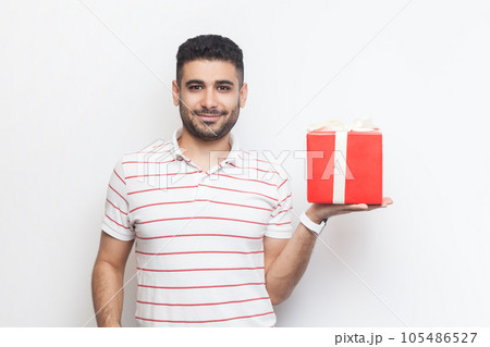Portrait of delighted cheerful handsome bearded man wearing striped t-shirt standing showing red wrapped present box, gift for birthday. Indoor studio shot isolated on gray background. 105486527