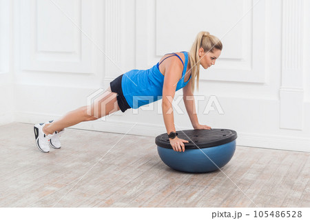 Full length portrait of concentrated woman in black shorts and blue top working in gym doing plank for abdominal muscles on bosu balance trainer, holding balance on fitness ball. Indoor studio shot. 105486528